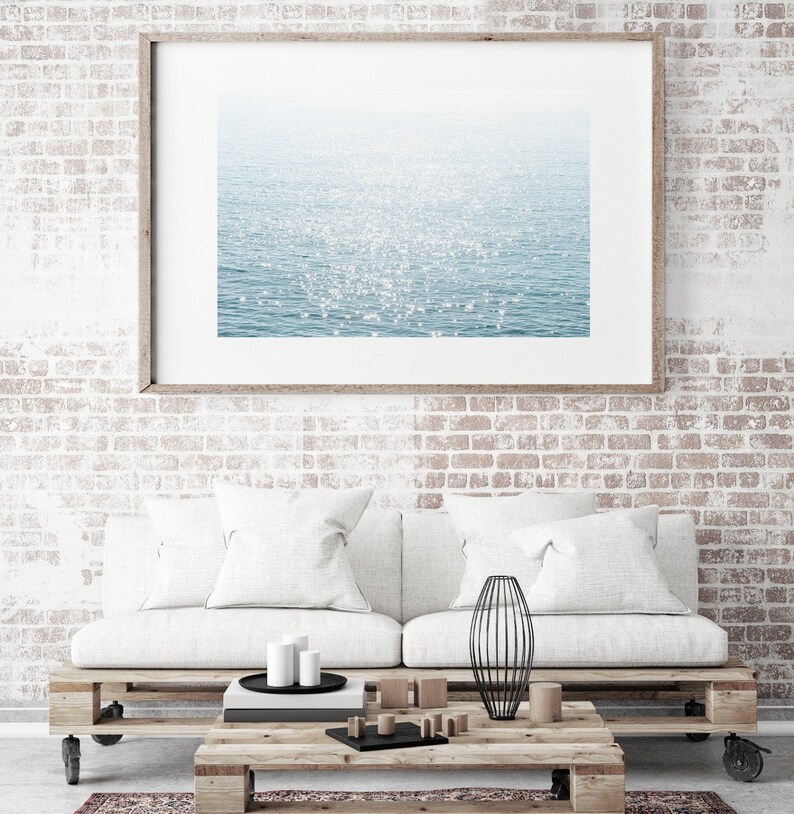 May include: A white couch with two white pillows sits in front of a brick wall. A framed photograph of a sparkling blue ocean hangs above the couch. A wooden coffee table with a black metal vase sits in front of the couch.