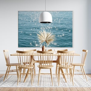 May include: A dining room table with six chairs, a light fixture, and a print of a blue ocean with sparkling light. The chairs are made of light wood and have a simple design. The table is made of light wood and has a rectangular shape. The print is hanging on the wall behind the table.
