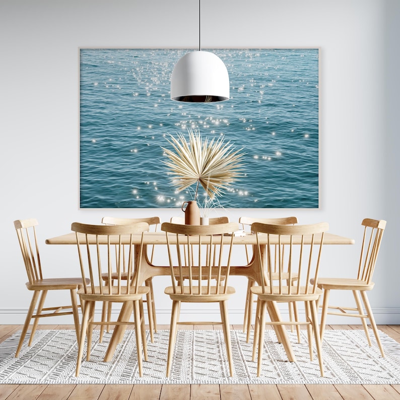 May include: A dining room table with six chairs, a light fixture, and a print of a blue ocean with sparkling light. The chairs are made of light wood and have a simple design. The table is made of light wood and has a rectangular shape. The print is hanging on the wall behind the table.