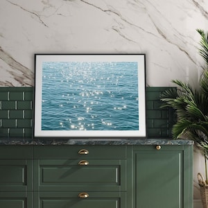 May include: A framed photograph of a blue ocean with sunlight reflecting off the water. The photograph is hanging on a wall above a green cabinet with gold handles.