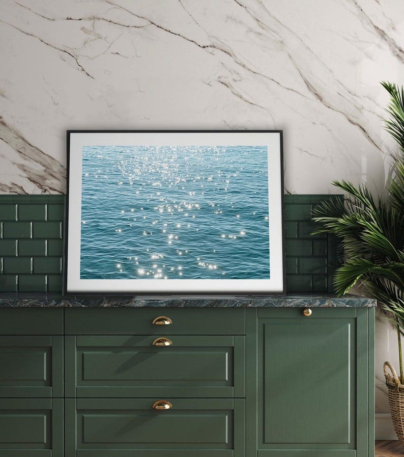 May include: A framed photograph of a blue ocean with sunlight reflecting off the water. The photograph is hanging on a wall above a green cabinet with gold handles.