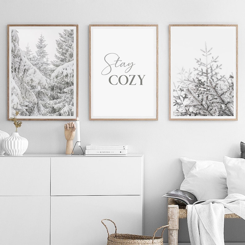 K&ouml;nnte beinhalten: Drei gerahmte Schwarzwei&szlig;drucke mit winterlichem Thema. Der mittlere Druck zeigt den Text "Stay Cozy" in grauer Schrift. Die Drucke werden an einer wei&szlig;en Wand &uuml;ber einer wei&szlig;en Kommode ausgestellt.