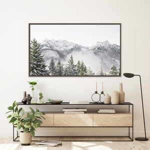 May include: A framed photograph of a snowy mountain range with a misty forest in the foreground. The photograph is hanging on a white wall above a wooden console table with a black metal frame. The console table has two drawers and is decorated with plants, candles, and a lamp.