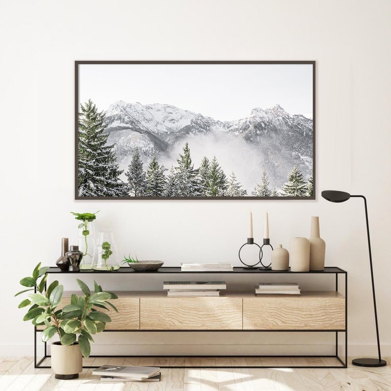May include: A framed photograph of a snowy mountain range with a misty forest in the foreground. The photograph is hanging on a white wall above a wooden console table with a black metal frame. The console table has two drawers and is decorated with plants, candles, and a lamp.