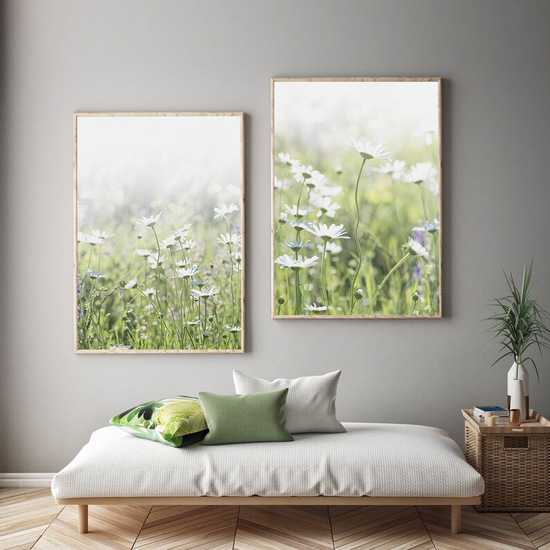 May include: Two framed prints of a field of white daisies with a soft, blurred background. The prints are hung on a light gray wall above a white couch with green pillows.