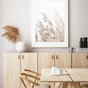 K&ouml;nnte beinhalten: Ein gerahmter Druck einer minimalistischen Naturszene mit hohen, federartigen Gr&auml;sern in Beige- und Wei&szlig;t&ouml;nen vor einem hellen Hintergrund. Der Druck h&auml;ngt an einer hellbraunen Wand mit einem Schrank und einem Tisch aus Holz darunter.