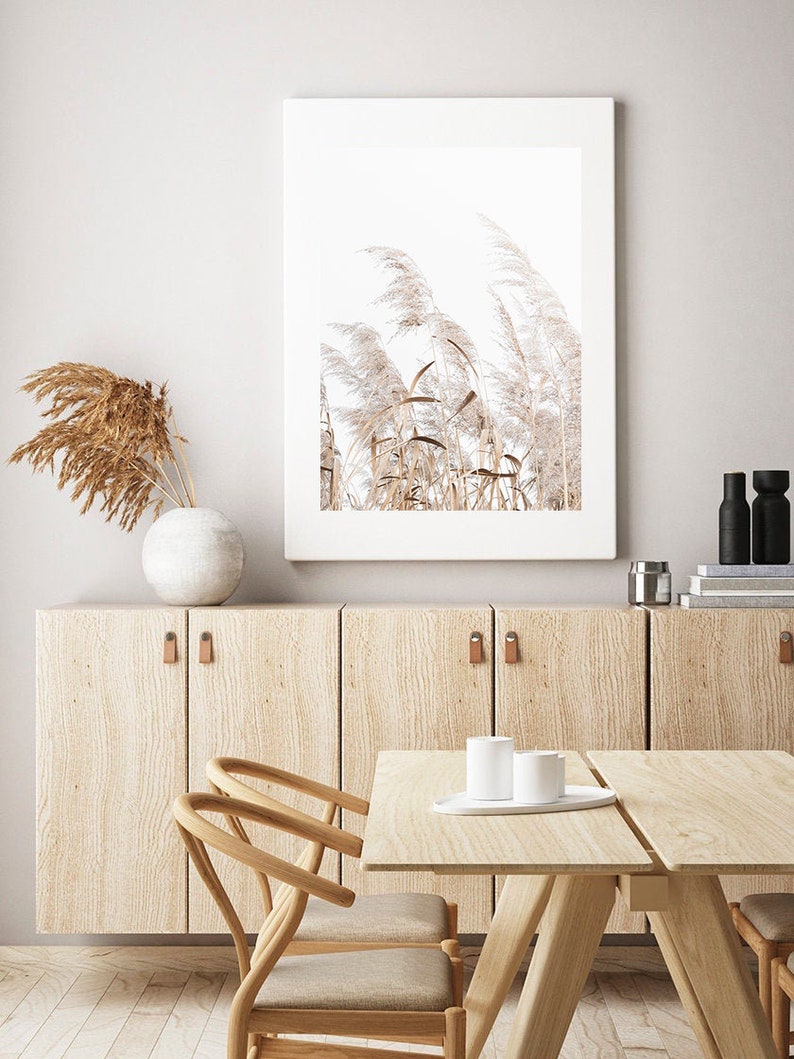 K&ouml;nnte beinhalten: Ein gerahmter Druck einer minimalistischen Naturszene mit hohen, federartigen Gr&auml;sern in Beige- und Wei&szlig;t&ouml;nen vor einem hellen Hintergrund. Der Druck h&auml;ngt an einer hellbraunen Wand mit einem Schrank und einem Tisch aus Holz darunter.