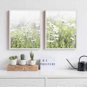 May include: Two framed prints of white daisies in a field. The prints are hung on a white wall above a white dresser with a wooden shelf, three potted succulents, a black watering can, and a white light-up sign that says "BE POSITIVE".