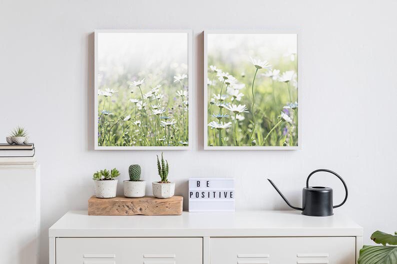 May include: Two framed prints of white daisies in a field. The prints are hung on a white wall above a white dresser with a wooden shelf, three potted succulents, a black watering can, and a white light-up sign that says "BE POSITIVE".