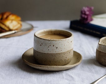 May include: A speckled brown and white ceramic mug filled with coffee sits on a white saucer. A book and a small ceramic creamer are in the background.