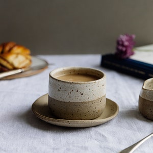 May include: A speckled brown and white ceramic mug filled with coffee sits on a white saucer. A book and a small ceramic creamer are in the background.