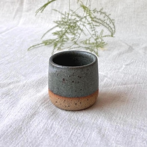 May include: A small, cylindrical ceramic cup featuring a two-tone design. The upper portion has a smooth, speckled dark grey glaze, while the lower portion displays a textured, natural tan clay finish with visible dark speckles throughout the surface.
