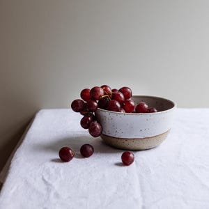 Può includere: Una ciotola di ceramica bianca con un design maculato, piena di uva rossa. La ciotola è appoggiata su un panno bianco, con qualche acino sparso. La ciotola ha un'estetica naturale e terrosa.