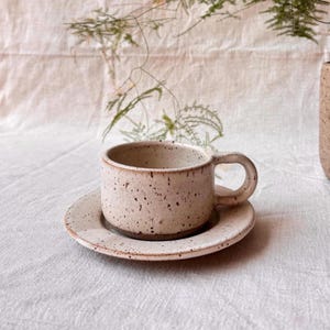 Könnte beinhalten: Ein Keramik-Tassen- und Untertassen-Set mit einer gesprenkelten, cremefarbenen Glasur und dunkelbraunen Punkten. Die Tasse hat einen breiten, zylindrischen Körper mit einem abgerundeten Henkel an der Seite, und die Untertasse ist passend gestaltet.