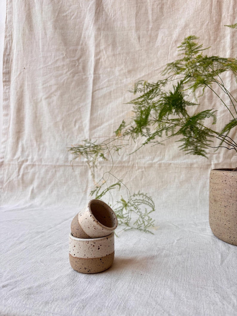 May include: Two small, cylindrical ceramic cups stacked on top of each other, featuring a speckled cream glaze on the upper half and a raw, textured brown clay base. A larger, matching speckled ceramic planter sits to the right.
