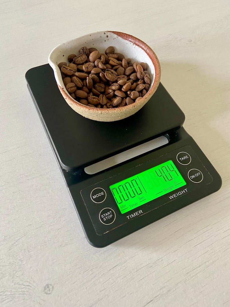 May include: A black digital kitchen scale with a green display showing "0.0001" and "404". The scale has a timer with buttons for "Start/Stop" and "Mode". A small white ceramic bowl filled with roasted coffee beans sits on the scale.