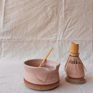 Pode incluir: Uma tigela de matcha de cerâmica rosa com uma colher de pau e um batedor de bambu. A tigela tem uma faixa marrom na base. O batedor é marrom claro e fica em uma base pequena e redonda. Em uma superfície branca texturizada.