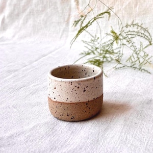 Könnte beinhalten: Ein kleiner, zylindrischer Keramikbecher mit zweifarbigem Design. Der obere Teil ist mit einer gesprenkelten, cremeweißen Glasur versehen, während der untere Teil aus einer strukturierten, unglasierten Basis aus braunem Ton besteht.
