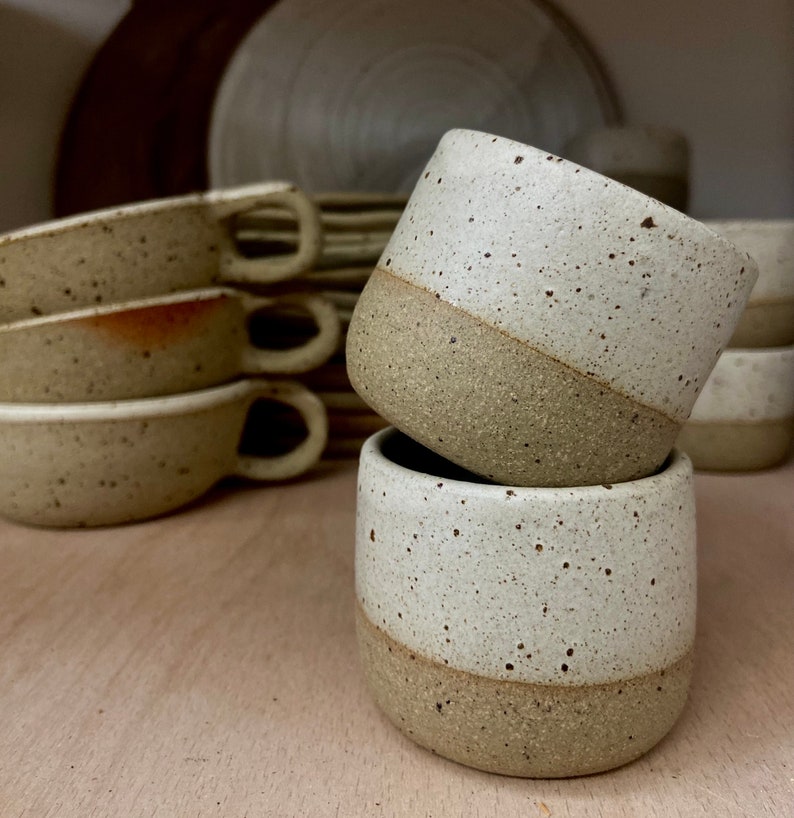 May include: Two stacked ceramic cups with a speckled cream glaze on the upper half and a solid tan base. The cups have a cylindrical shape with no handles. In the background, several other ceramic cups with handles are visible in a stack.