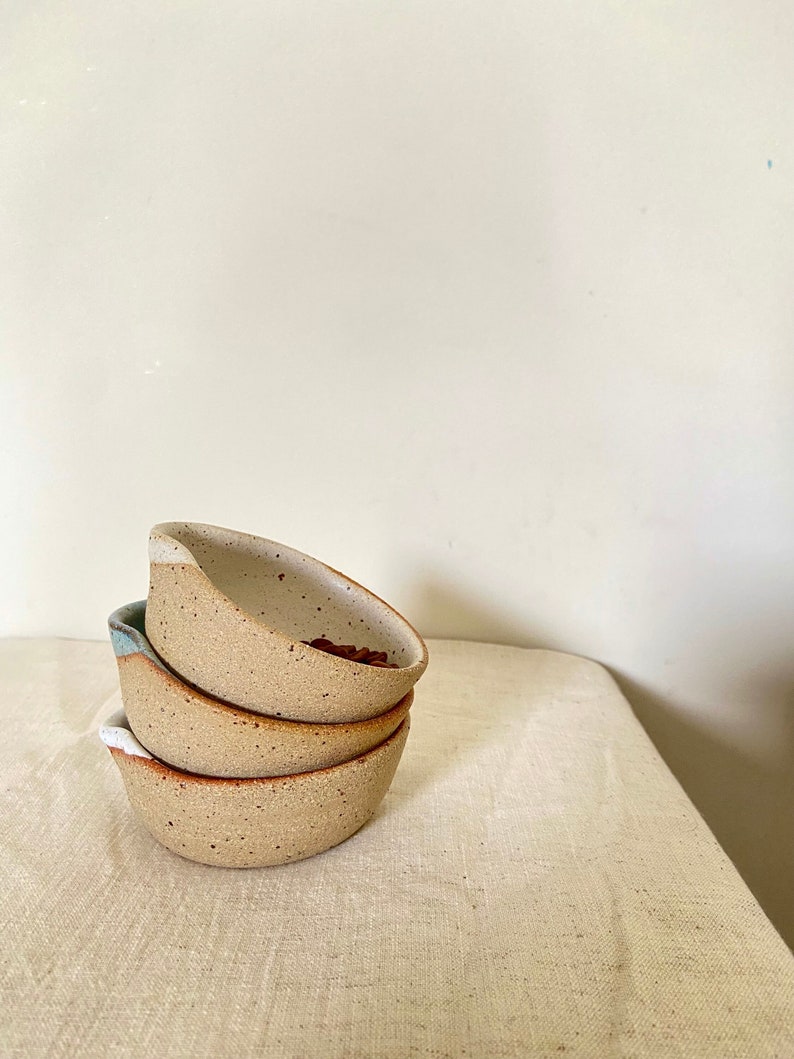 May include: A stack of three small, speckled ceramic bowls with a white rim and a light blue glaze. The bowls are nested together on a white linen tablecloth.