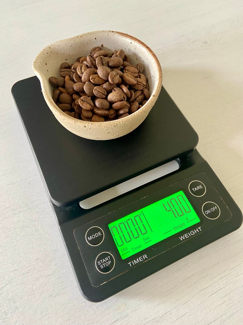 May include: A black digital kitchen scale with a green display showing a weight of 400 grams. A white ceramic bowl filled with roasted coffee beans sits on the scale.