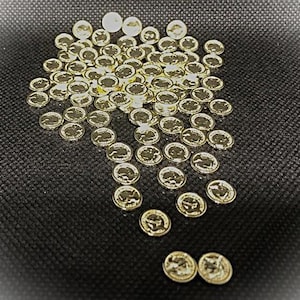 Puede incluir: Una colección de pequeñas monedas doradas dispersas sobre una superficie oscura y texturizada. Las monedas parecen ser réplicas en miniatura, posiblemente para manualidades o coleccionismo. Los detalles de las monedas son visibles.