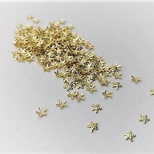 May include: A scattering of small, gold-coloured star-shaped embellishments on a white surface. The stars have a textured appearance and are clustered together, with some individual stars spread out.