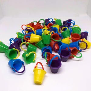 May include: A pile of miniature toy buckets in a variety of colours: red, yellow, green, blue, and purple. Each bucket has a matching handle. The buckets are scattered on a white surface.