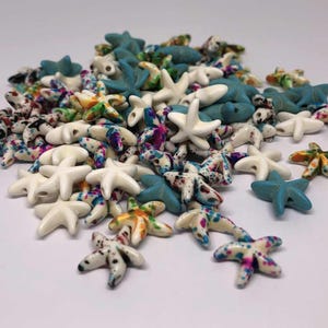 May include: A pile of starfish-shaped beads in various colours. The beads are white, turquoise, and multi-coloured with speckled patterns. Some beads have holes for stringing. Suitable for jewellery making or craft projects.