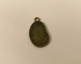 May include: A bronze-coloured metal charm shaped like a melting clock face with Roman numeral markings and a single hand pointing to the 3. The charm has a loop at the top for attaching to a necklace or bracelet.
