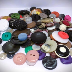 Peut inclure: Une grande collection de boutons de couture assortis de formes, tailles et couleurs variées, notamment noir, blanc, rose, vert, violet et rouge. Les boutons présentent différentes textures, motifs et configurations de trous, éparpillés en tas.