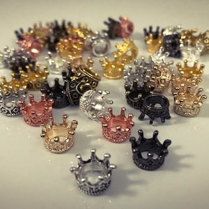 May include: A collection of small, gold, silver, and black crown-shaped metal beads. The beads are scattered on a white surface.