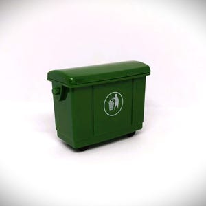 May include: A miniature green wheelie bin with a rounded lid. It features a white circular graphic of a person disposing of waste. The bin is on wheels and has a handle on the side. It is a model.