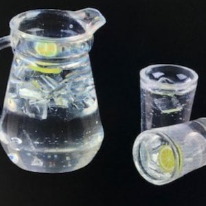 May include: A clear glass jug filled with ice and lemon slices, next to two clear glass tumblers, one upright and one lying down, also filled with ice and lemon slices.
