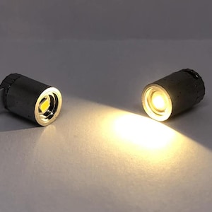 May include: Two small, cylindrical LED lights with a bright, warm glow. One has a white tab attached. The lights are silver and gray, casting a bright beam on a white surface.