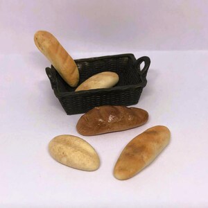 May include: A miniature bread basket with several small loaves of bread. The basket is dark green and contains two loaves. Three additional loaves are arranged around the basket. The bread is various shades of brown and tan.