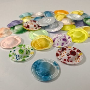 Peut inclure: Un assortiment de petits boutons recouverts de tissu, aux couleurs vives : jaune, vert, bleu, rose et violet. Certains boutons présentent des motifs floraux, d'autres sont unis. Les boutons sont disposés sur une surface blanche.