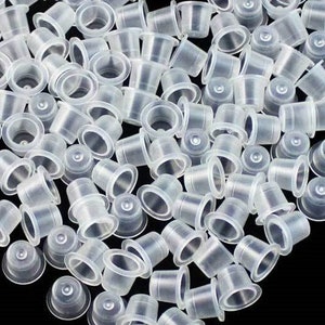 May include: A large group of clear plastic tattoo ink cups. The cups are stacked and scattered on a black surface.