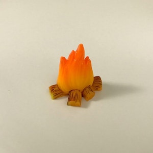Puede incluir: Una fogata en miniatura de color naranja y amarillo con troncos marrones. La fogata está hecha de plástico y mide aproximadamente 5 cm de alto.
