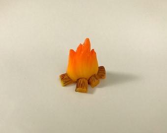 Könnte beinhalten: Ein Miniatur-Lagerfeuer in Orange und Gelb mit braunen Holzscheiten. Das Lagerfeuer ist aus Plastik und etwa 5 cm hoch.