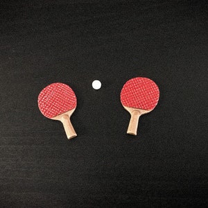Puede incluir: Dos palas de ping pong rojas y negras con mangos de madera sobre una superficie negra con una pelota de ping pong blanca en el centro.