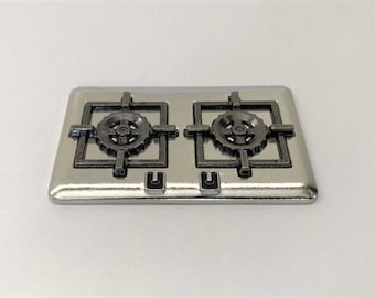 Könnte beinhalten: Ein silberfarbenes Kochfeld mit zwei schwarzen Brennergittern. Jedes Gitter hat ein kreisförmiges Design in der Mitte.