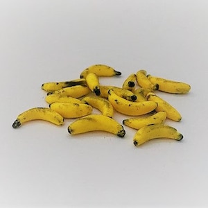 Op de afbeelding: Een cluster van kleine, gele bananen met donkere uiteinden, gerangschikt op een witte ondergrond. De bananen hebben een licht gebogen vorm en lijken rijp. De afbeelding is goed belicht, waardoor de textuur en kleur van het fruit worden benadrukt.
