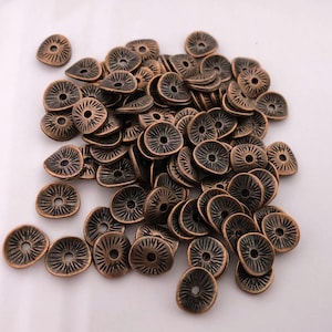 Może przedstawiać: Kolekcja metalowych koralików w kolorze brązu. Każdy koralik ma okrągły wzór z centralnym otworem i promienistymi liniami. Koraliki mają nieregularny kształt i wydają się być wykonane z trwałego metalu.