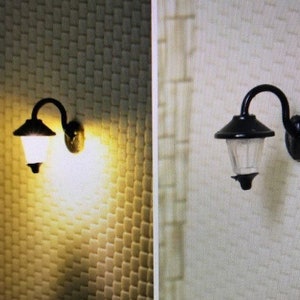 Può includere: Due applique da parete nere con paralumi in vetro trasparente. L'applique a sinistra è accesa e illumina la parete.