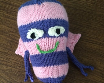 Puede incluir: Un juguete de rana de punto con rayas rosas y moradas, ojos blancos con pupilas negras y una boca sonriente verde. La rana tiene patas largas y delgadas y una pequeña ala a cada lado.