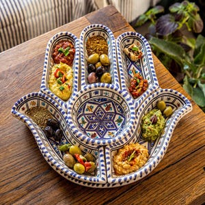 May include: A hand-shaped ceramic serving platter featuring a white base with intricate blue and multi-coloured geometric patterns. The dish is divided into several compartments, each filled with various dips, olives, and herbs for serving.