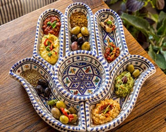 Puede incluir: Una bandeja de cerámica con forma de mano que presenta una base blanca con intrincados patrones geométricos en azul y varios colores. El plato está dividido en varios compartimentos, cada uno lleno de diversos dips, aceitunas y hierbas.
