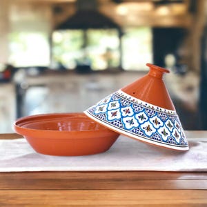 May include: A terracotta tagine with a white and blue patterned lid. The lid has a small hole in the top. The tagine is sitting on a wooden table.