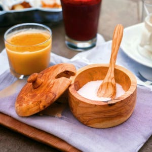 Puede incluir: Un salero de madera con tapa y cuchara a juego, lleno de sal blanca. El salero y la cuchara son de madera de color marrón claro. Un vaso de zumo de naranja y un vaso de zumo rojo están en el fondo.